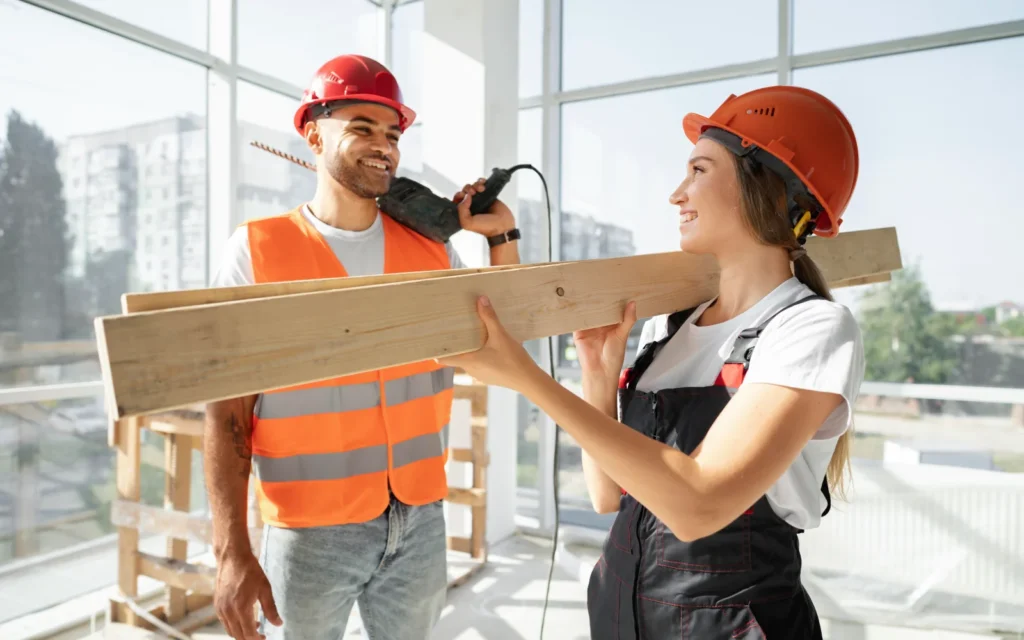 Dos trabajadores de la construcción, un hombre y una mujer, sonríen mientras sostienen madera en un ambiente luminoso y moderno. Ambos llevan cascos de seguridad y ropa adecuada para el trabajo. La imagen destaca la colaboración en proyectos de construcción.