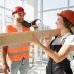 Dos trabajadores de la construcción, un hombre y una mujer, sonríen mientras sostienen madera en un ambiente luminoso y moderno. Ambos llevan cascos de seguridad y ropa adecuada para el trabajo. La imagen destaca la colaboración en proyectos de construcción.