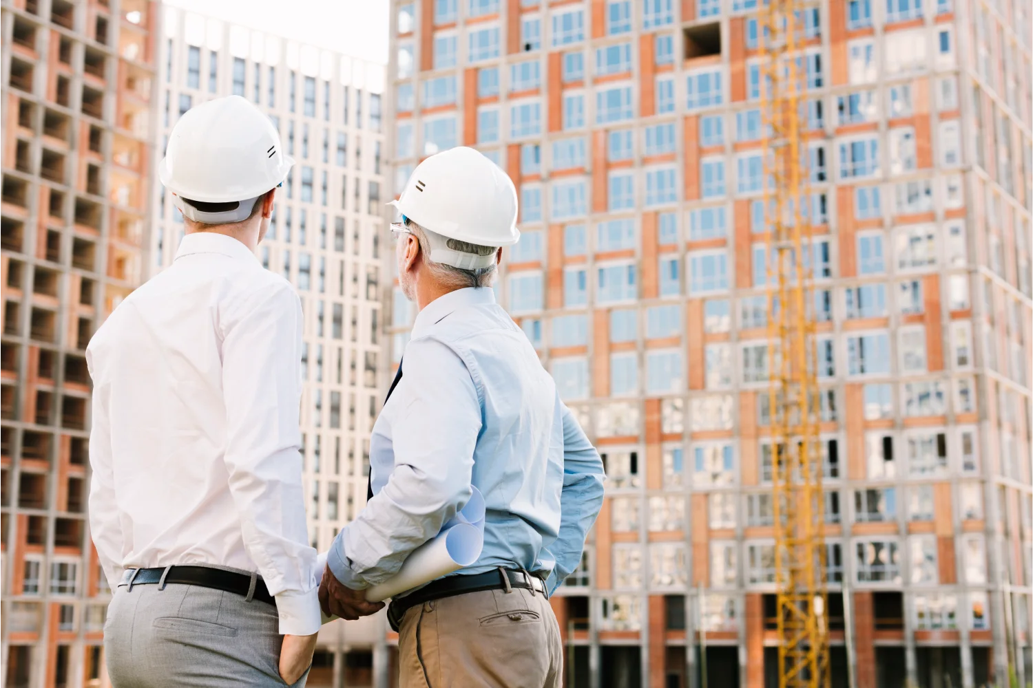 Ingenieros observando la construcción de un edificio moderno, con planos en mano y cascos de seguridad, en un sitio de obra.
