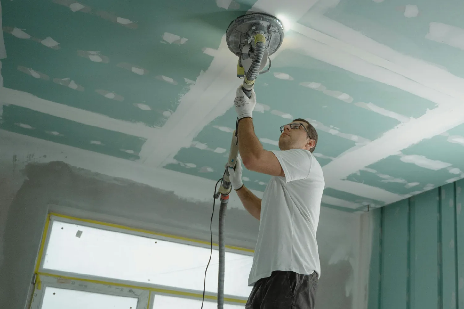 Hombre realizando trabajos de construcción, utilizando una herramienta eléctrica para lijar el techo en un ambiente de renovación. Se observan paneles de yeso y una ventana con luz natural.