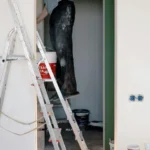 Trabajador en una escalera pintando una pared interior, con cubos de pintura y herramientas de trabajo visibles en el fondo.