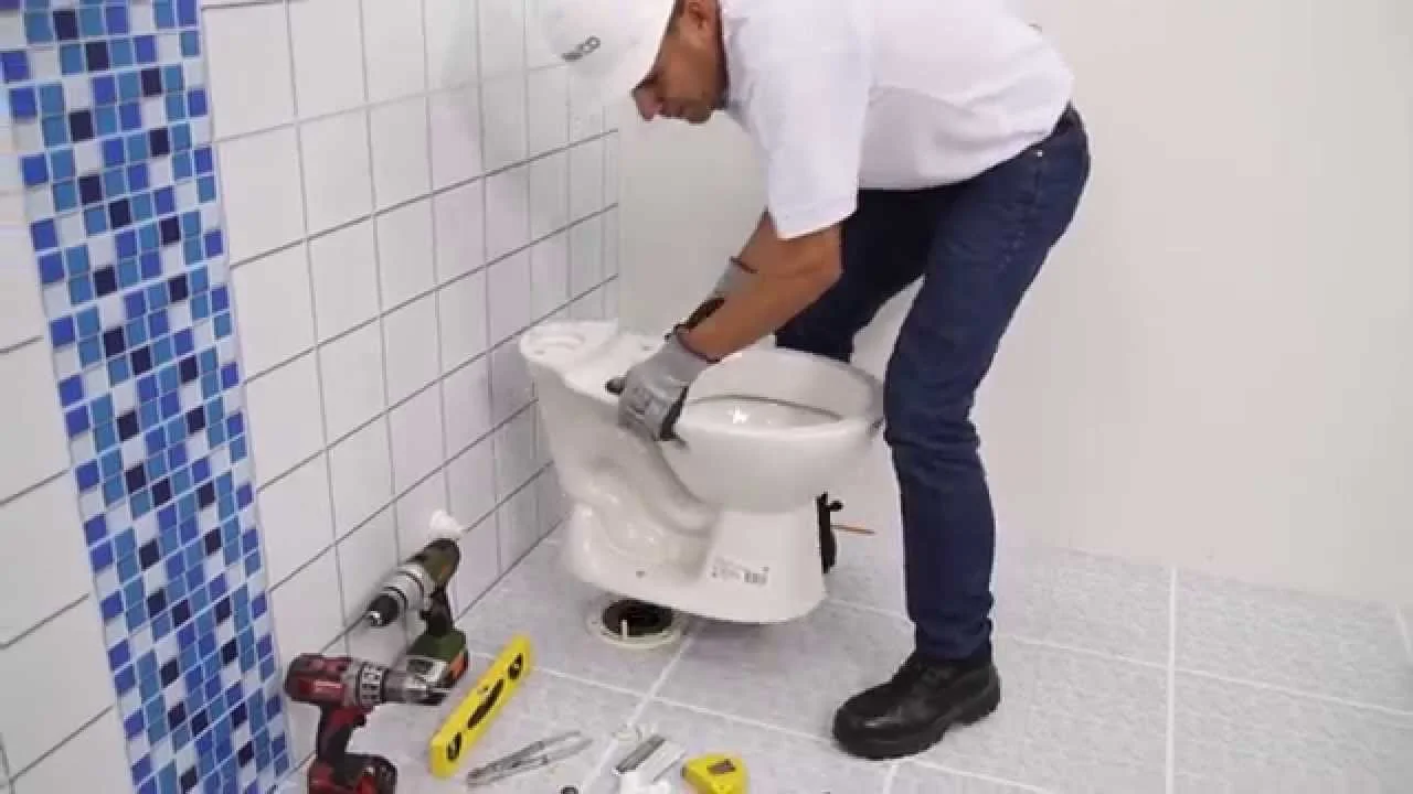 Instalador de inodoro en proceso de colocación, rodeado de herramientas como taladro y nivel, en un baño con azulejos blancos y azules.