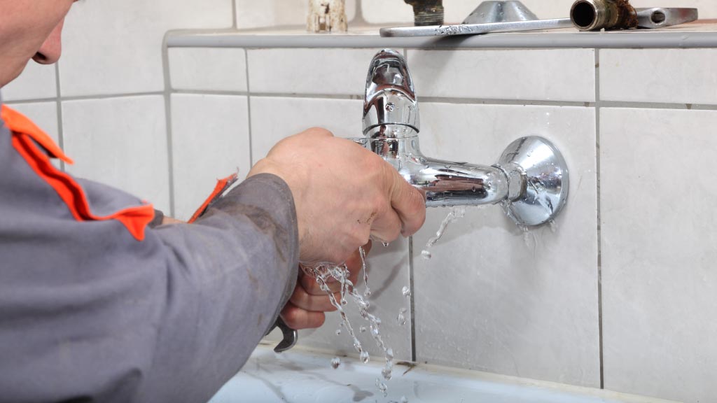 Plomero reparando un grifo en un baño, con agua fluyendo de la llave. Herramientas visibles en el fondo, mostrando un trabajo de fontanería en progreso.
