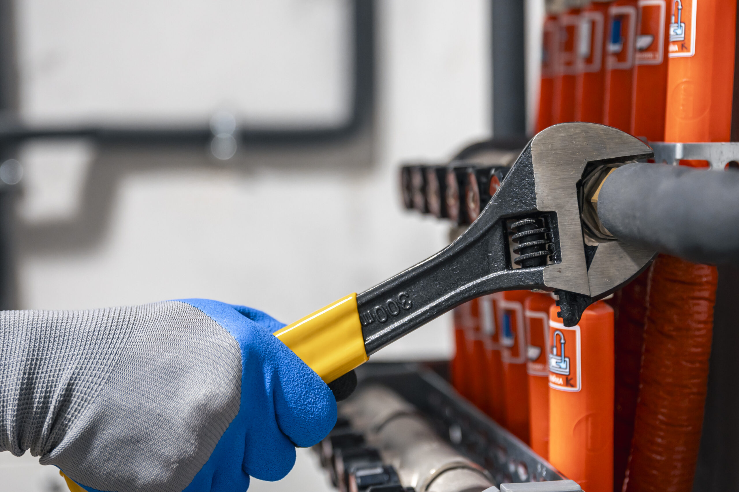 Llave ajustable negra en uso para apretar una tubería en un entorno industrial, con fondo de herramientas y tuberías de colores.